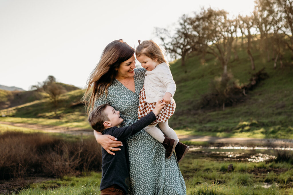 A mommy and me photography session with Jenn Chen Photography. A mom is holding her daughter while her son is hugging them both. All three are looking at each other smiling. The Walnut Creek back ground is a beautiful hilly park with a warm glowing sun.