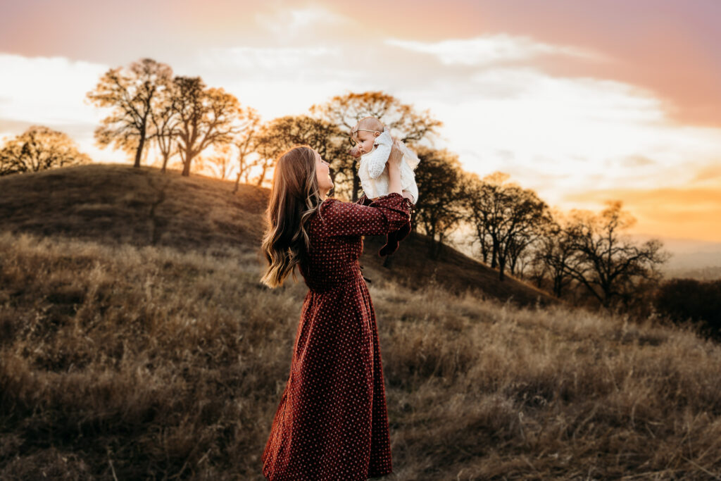 a mom enjoying her east bay photography session as she lifts her baby up and smiles at her. The sun is setting behind them and turning the sky a pinkish orange.