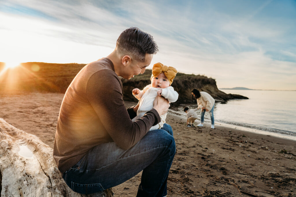 A family of 4 during their photography session in Hercules, CA. The dad and baby sit on a log, and the mom and toddler are in the background collecting shells on the beach.