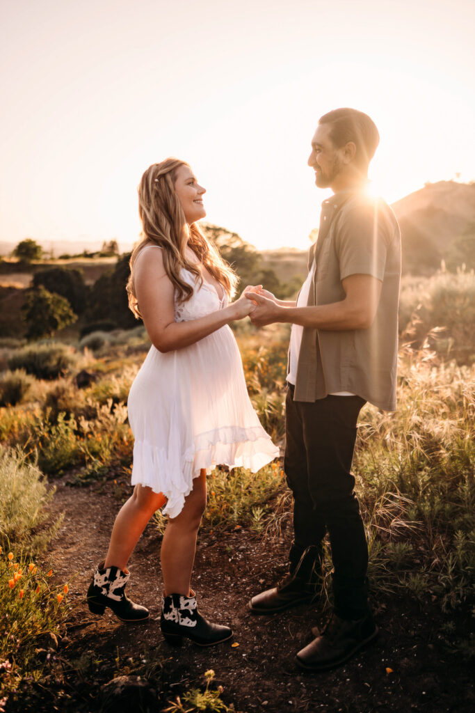 The sun shines behind a couple holding hands. The woman is pregnant and wearing a white maternity dress.