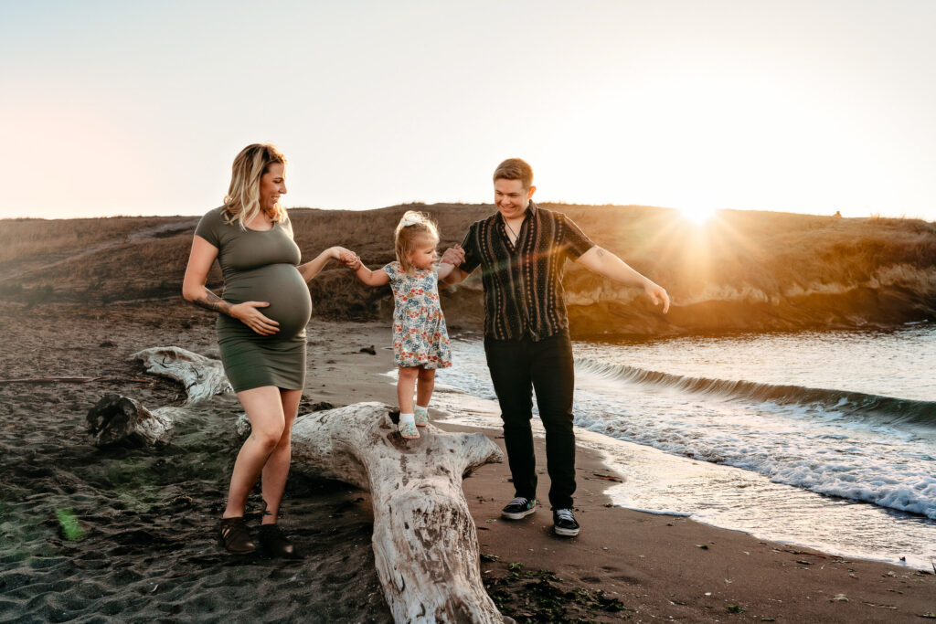 a family of 3. a pregnant mama wears a form fitting maternity dress and holds her toddlers hand, smiling. You can see the Martinez CA beach behind them.