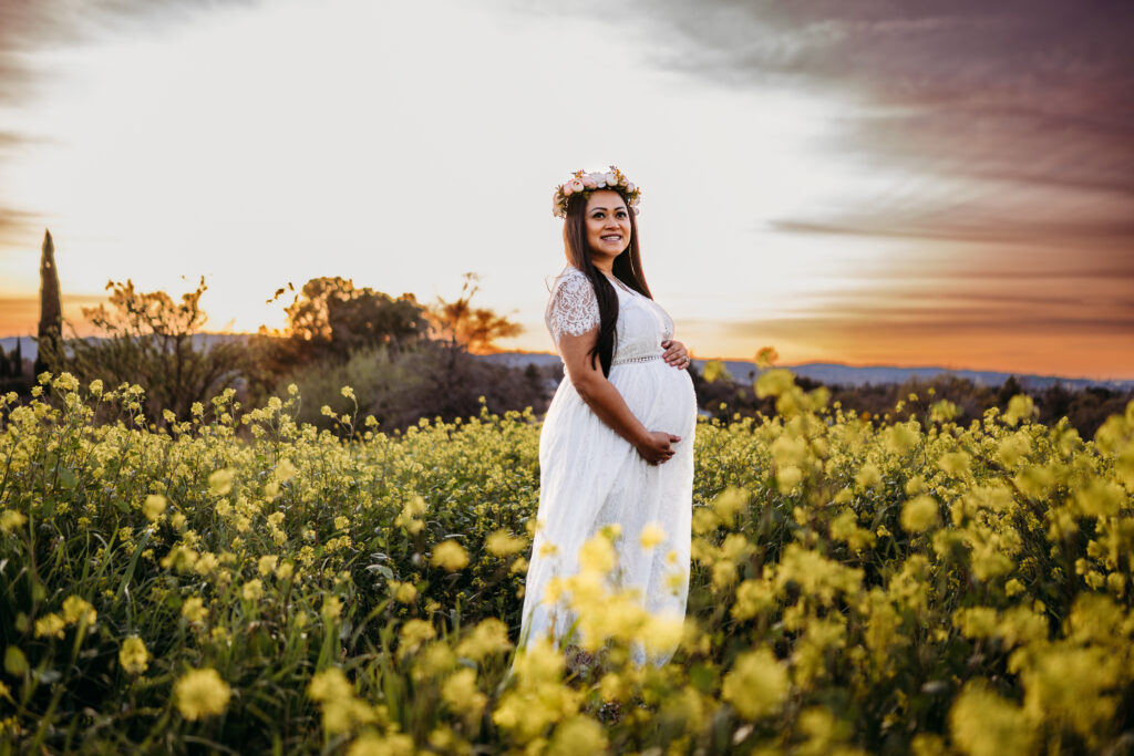 a maternity session in walnut creek with a mom wearing a flower crown. she stand smiling in a field of wildflowers under a colorful sunset