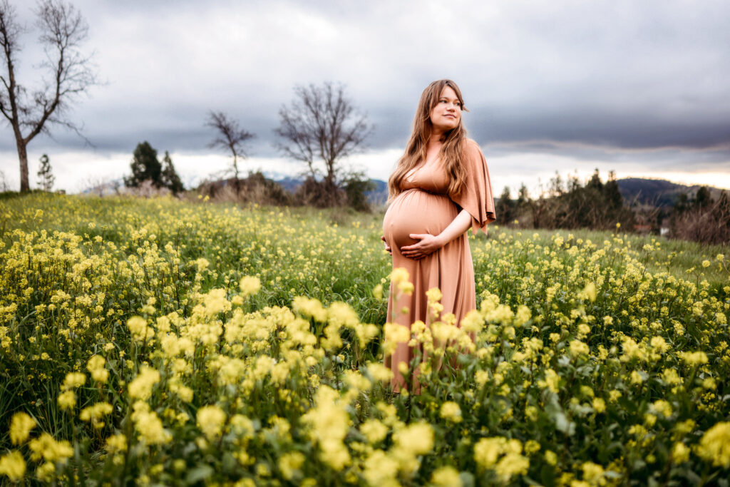 the same mom in a salmon maternity dress looks out over the huge field of mustard flowers. her hands are on her belly.