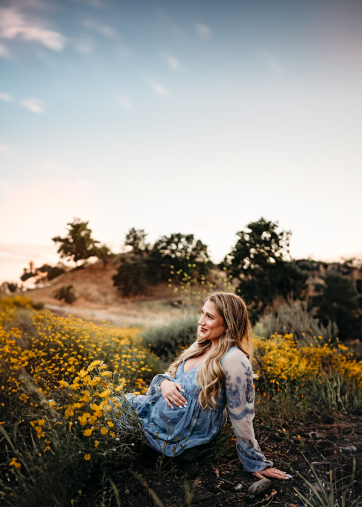 mom in blue maternity dress sit on top of a hill overlooking fields of wildflowers below