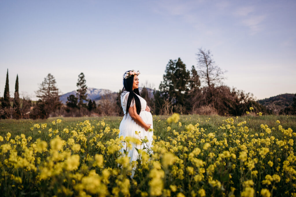 a pregnant mom in a white maternity dress looks off into a giant field of wild flowers. mount diablo in in the distance