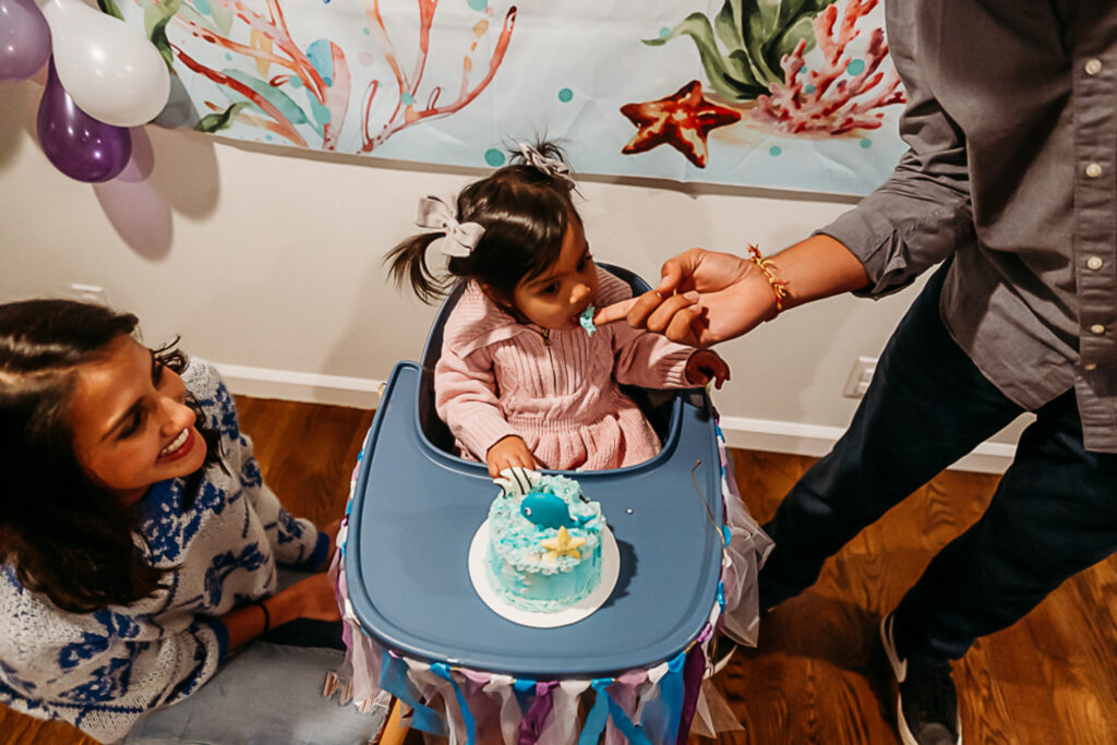 dad feeds baby a bit of frosting off of her birthday cake. mom is next to her smiling