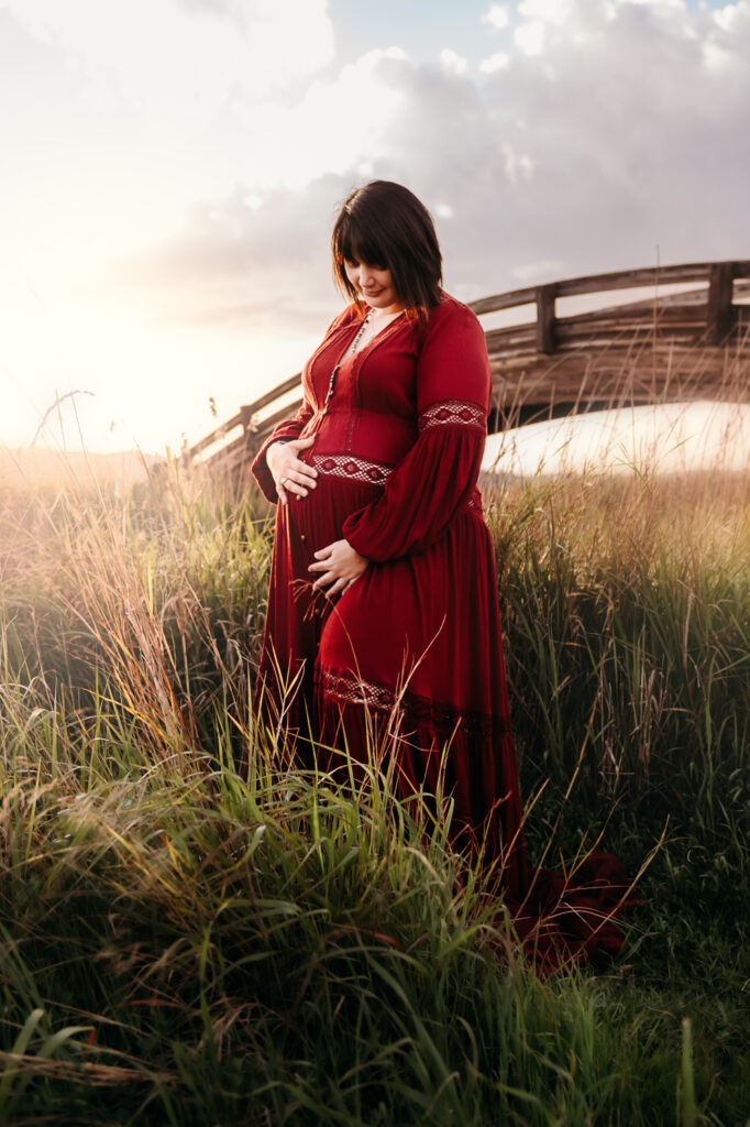 mom in red maternity dress stands in front of a arch bridge 