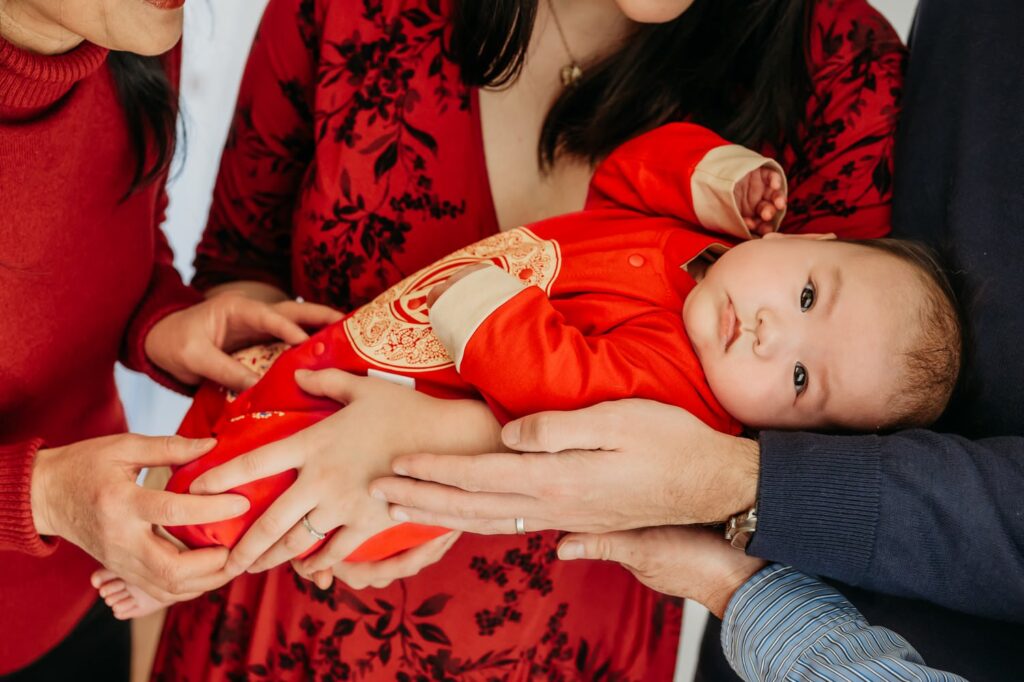 Close of of a family during their 100 days studio session. The baby wears a traditional red outfit and you can see her families hands all around her.