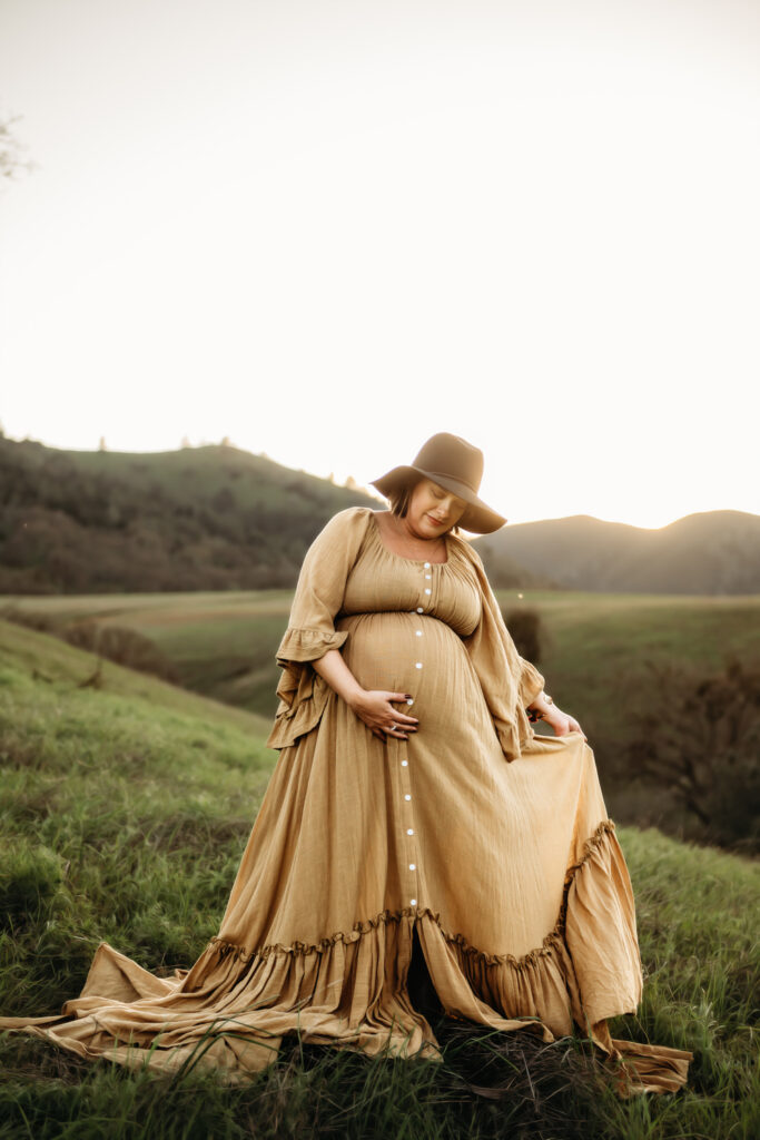 pregnant mama looks down smiling at her baby bump. she holds her flowing maternity dress in one hand and her belly with the other.