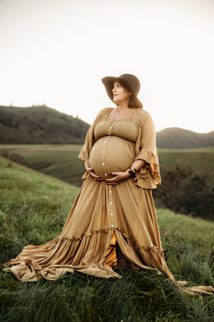 close up of a pregnant mama. she has a long mustard colored maternity dress. mt diablo is in the distance behind her.