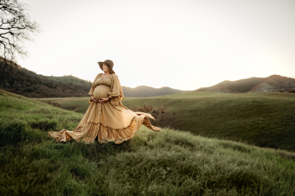 woman in flowing mustard colored maternity dress and a hat. her hands are on her belly and she is looking towards the mountains