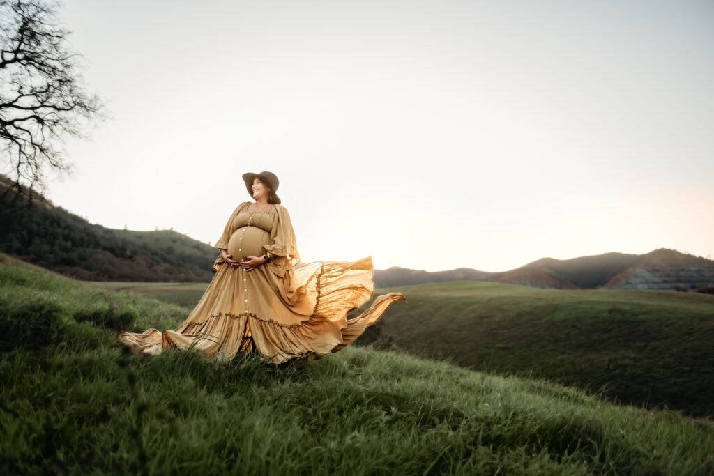 pulled back shot of a mama wearing a dramatic maternity dress blowing in the wind. the sun sets behind mt diablo in the distance behind her.