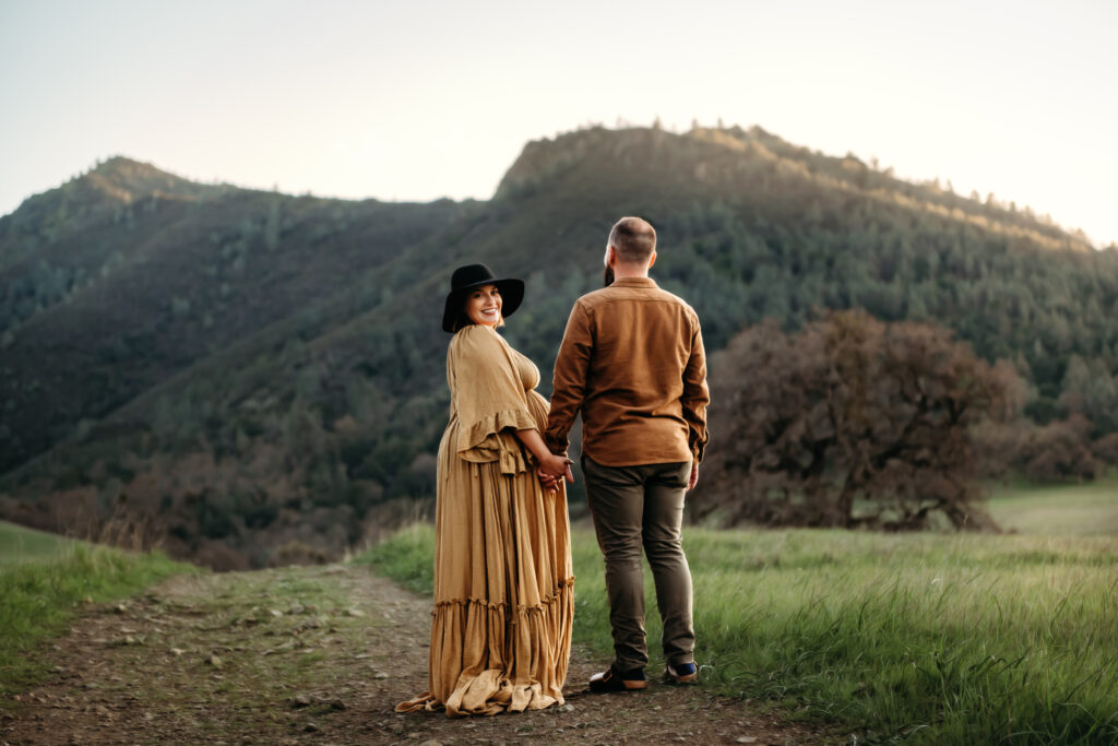 pregnant woman looks over her shoulder towards the camera. she is holding hands with her husband. they are walking down a trail at the base of mt diablo.