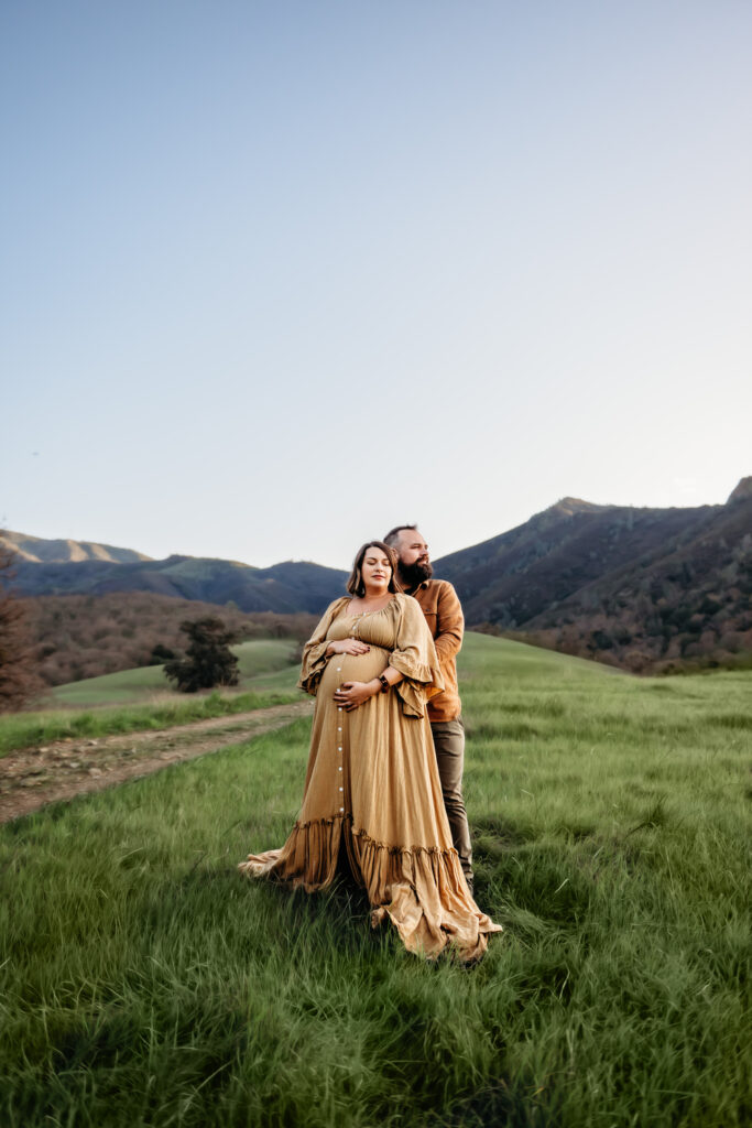 husband hugs his pregnant wife from behind. she has her eyes closed and her hands on her baby bump. mt diablo rises behind them