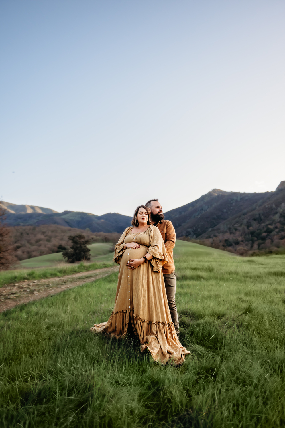 a maternity session at the base of mount diablo featuring a pregnant mama in a mustard colored maternity dress and her husband. the husband holds her from behind and looks off camera while the mama closes her eyes and looks calm.