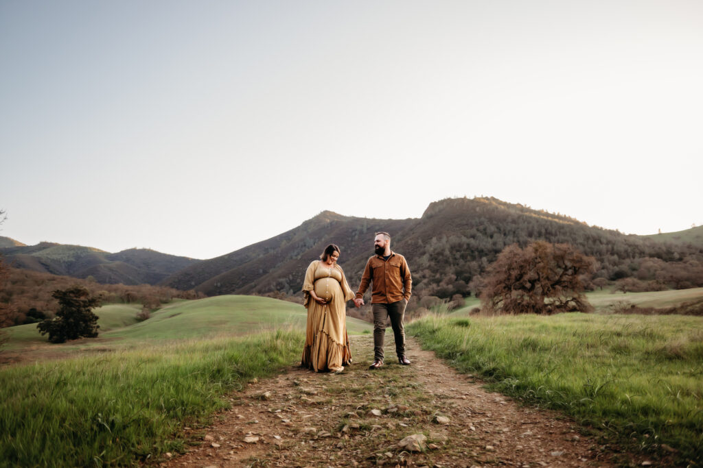 pregnant mama laughs and her husband looks at her. they are holding hands and walking down one of the trails at the base of mt diablo.