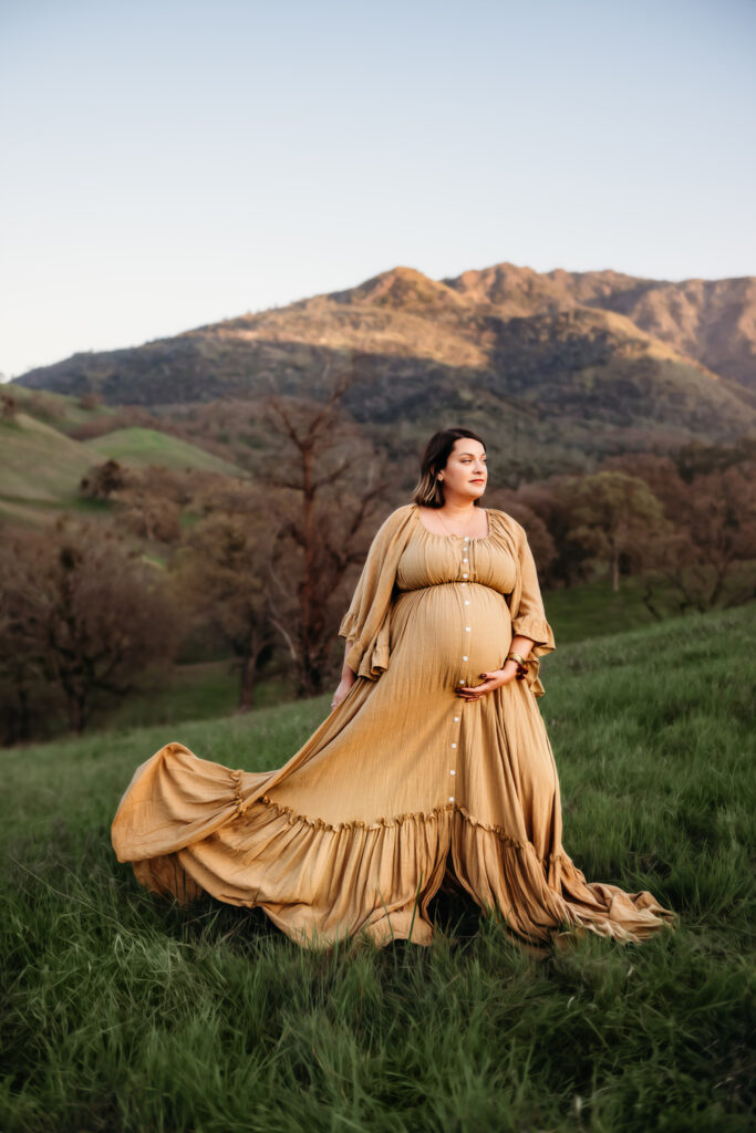 pregnant mama in a yellow maternity dress. the wind softly blows her dress as she holds her belly looking off camera.