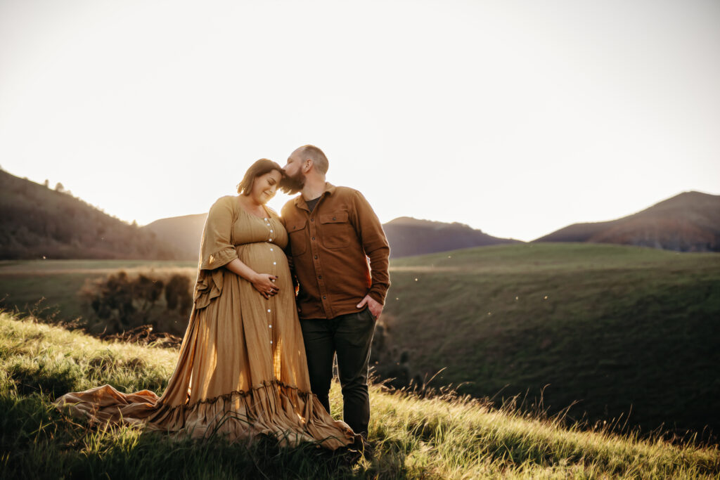 husband kisses wifes temple as she looks down at her pregnant belly smiling.