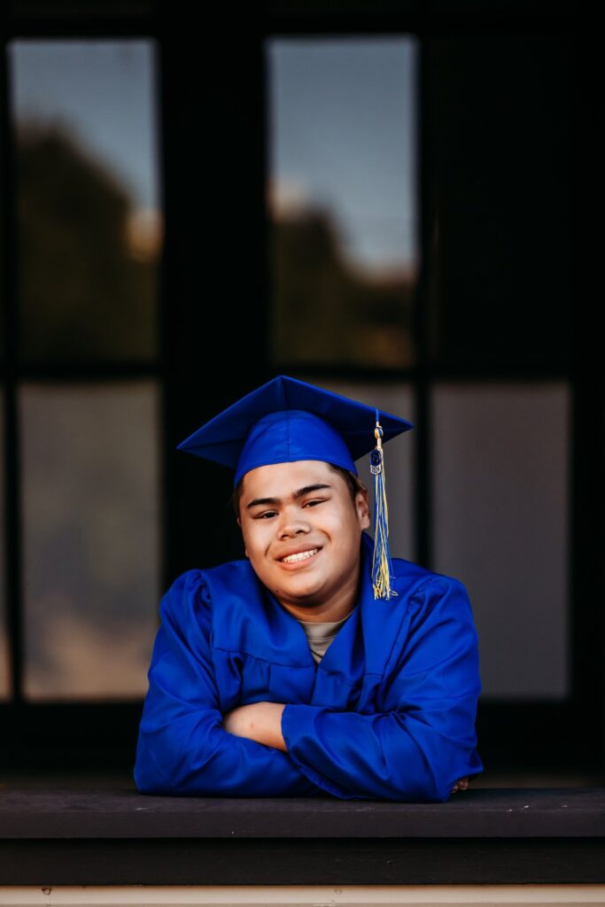 Close up of senior boy with dark background. He is wearing his blue cap and gown.