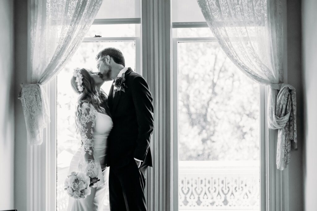 Classic black and white portrait of bride and groom in front of a window with lace curtains