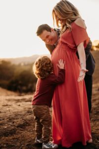 dad laughs as he watches his son kiss his wifes pregnant belly. he wears a pink maternity dress. the sun sets behind them