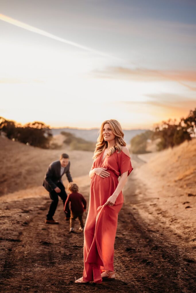 maternity photos at old borges ranch in walnut creek. mama wears a pink maternity dress and her family is in the hills behind her.