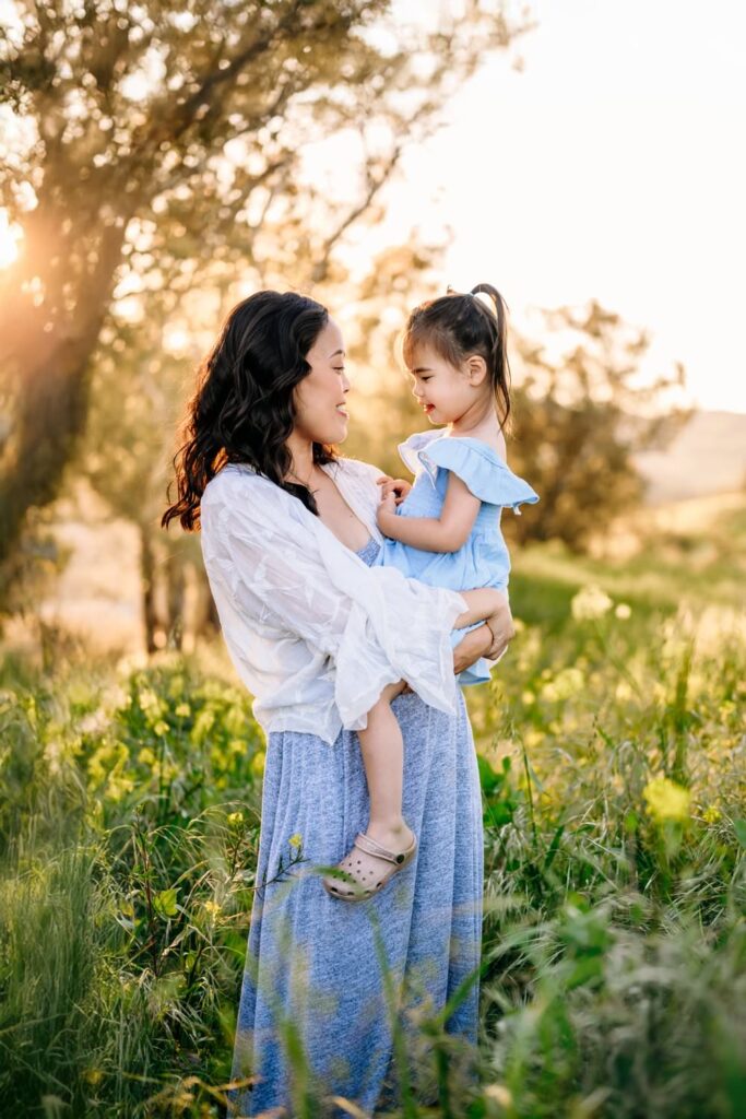 mom holding her toddler in a field of flowers ass the sun sets behind them
