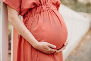 close up of a pregnant belly. mom wears a pink maternity dress. you see the white fence from old borgess ranch's goat farm behind her