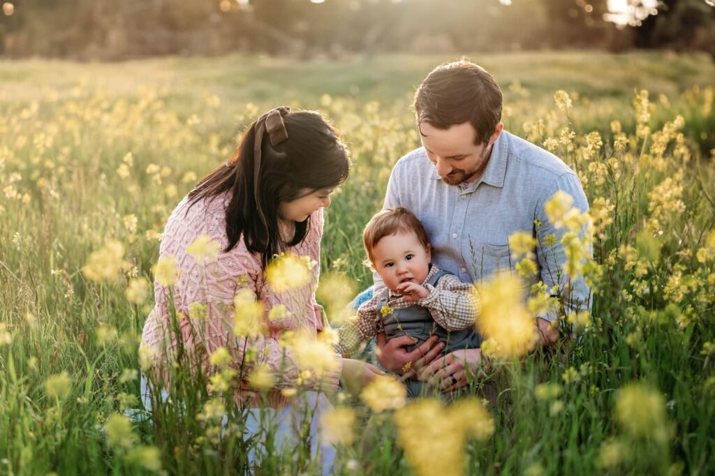 toddler looks at camera and parents look down at him. they are sitting in a huge field of wildflowers