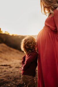 little boy kisses his moms belly as the sun sets behind them. mom smiles down at him