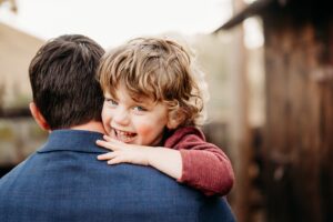 little boy laughs at the camera, looking over his dads shoulder