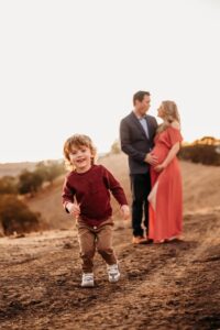 mom and dad are posing for maternity photos behind a running, smiling toddler