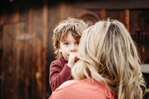 toddler sucks his thumb and looks over his moms shoulder at the camera during their photoshoot