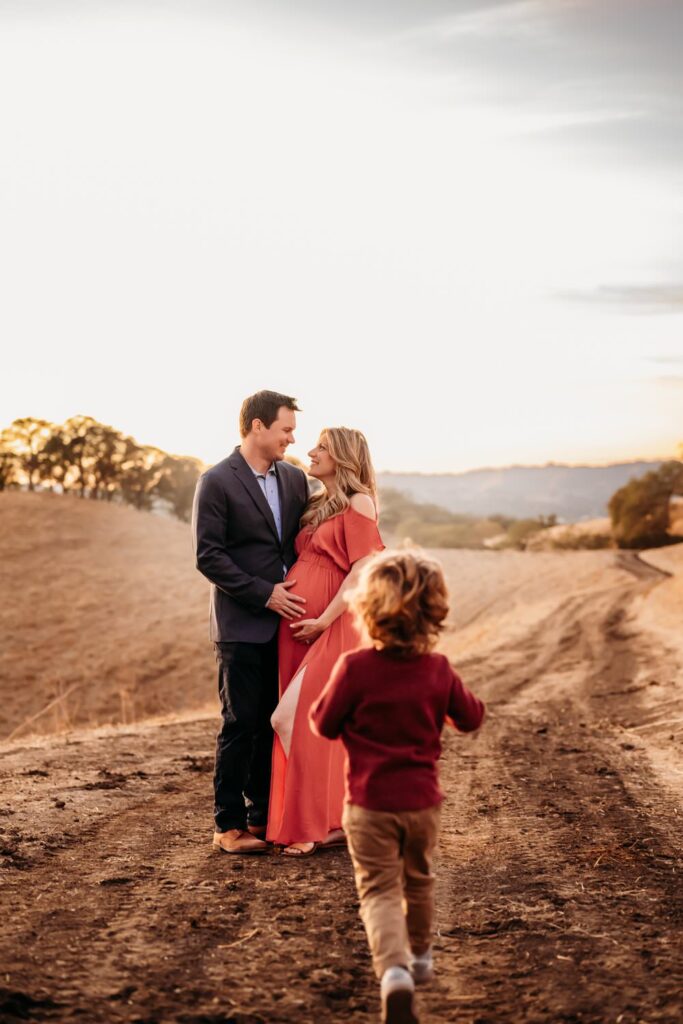 toddler runs toward his parents. dads hand is on moms pregnant belly