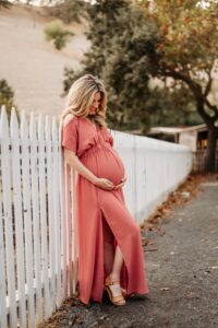 pregnant mom poses for maternity photos. she leans against a white fence looking down at per belly