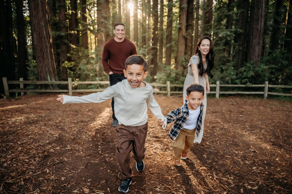 2 boys run towards camera holding hands with their mom holding their hand behind them. the sun is shining through the redwoods creating beautiful sun rays