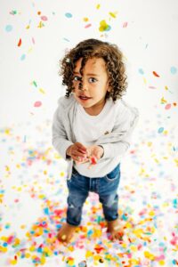 curly haired boy looks up at camera. he stands in a pile of multi colored confetti