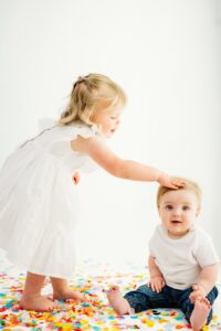 sister pats baby brothers head as he smiles at camera