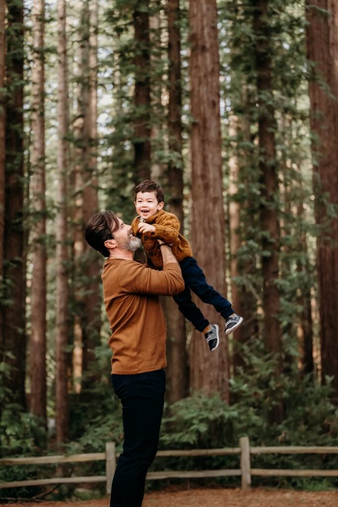 Parent lifting their young child into the air among tall redwood trees during an outdoor family photography session in the East Bay.