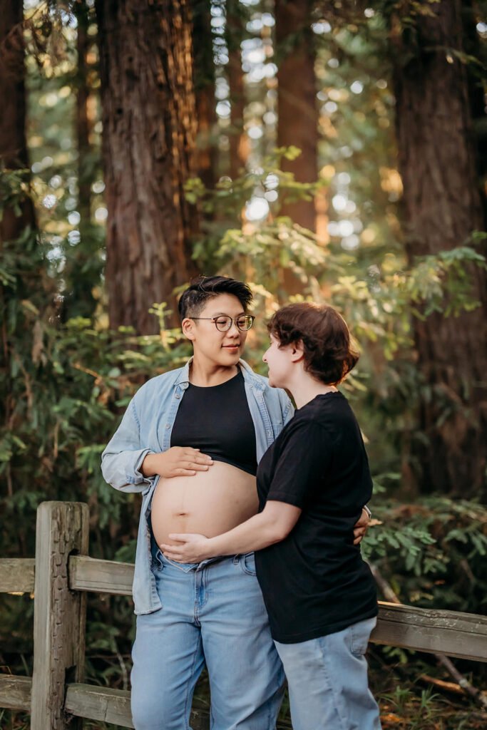 pregnant mom looks at her wife as the sun peeks through the redwoods behind them