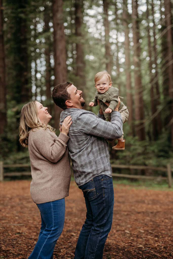 little boy looks at the camera as his dad lifts him and mom looks laughing