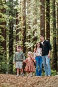 siblings walk through forest holding hands. their parents are behind them watching and smiling
