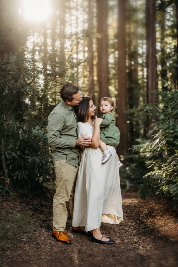 Parents standing together in a wooded trail holding their toddler close, sharing a quiet family moment during a Lamorinda photo session.