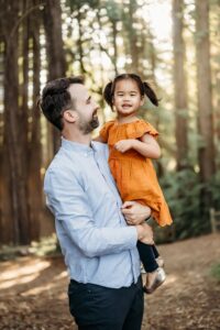 little girl in her dads arms. redwoods are behind them