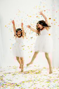 2 sisters in white dresses jump and dance in rainbow colored confetti
