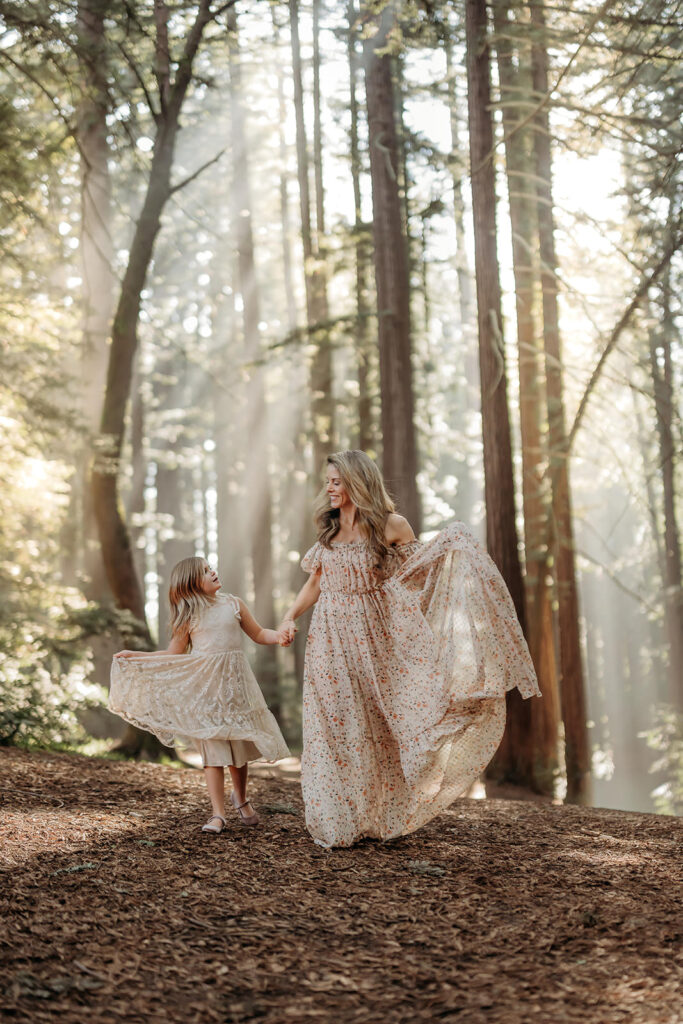 mom and daughter hold hands walking through a redwood forest swishing their dresses