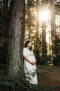 a pregnant mom leans against a redwood tree smiling