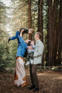 mom lifts her toddler while dad and baby watch. they are in a sunny redwood forest in oakland