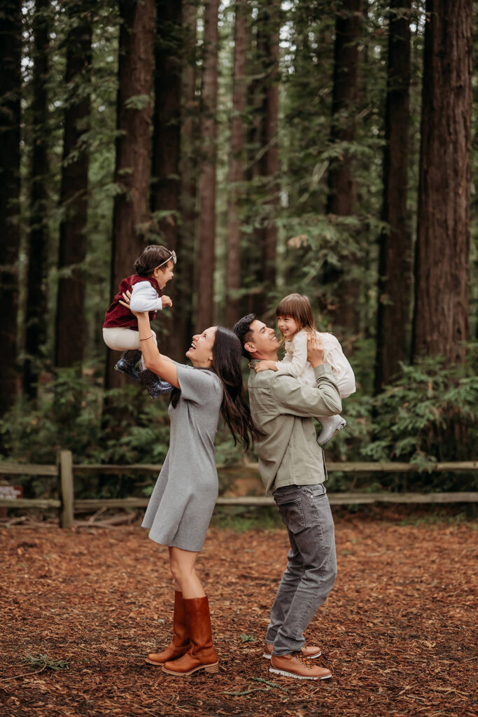 Parents lifting their children into the air while playing together in a redwood forest during a candid family photo session.