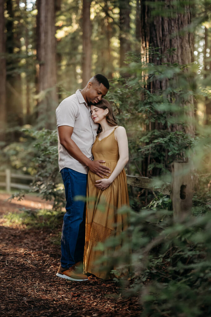 pregnant mama in mustard maternity dress with her husband. they are nestled in a redwood grove